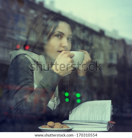 Young woman drinking coffee and reading book sitting indoor in urban cafe. Cafe city lifestyle. Casual portrait of teenager girl. Toned.