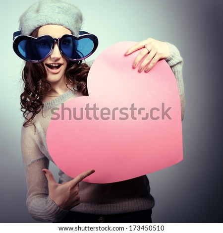 Cute attractive fashion young girl posing with funny big love glasses and pointing at pink heart, toned