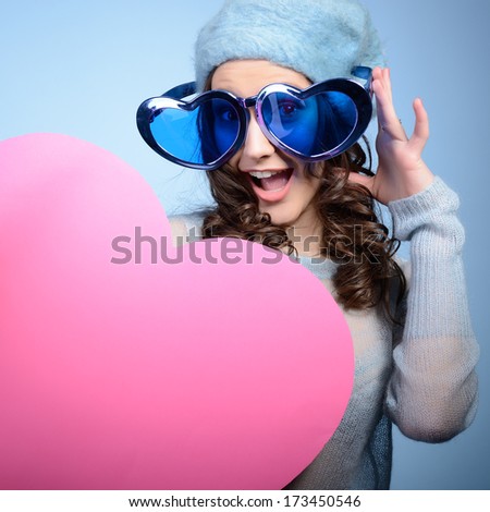 Cute attractive fashion young girl posing with funny big love glasses and pink heart over blue background Cute attractive fashion young girl posing with funny big love glasses and pink heart over blue background