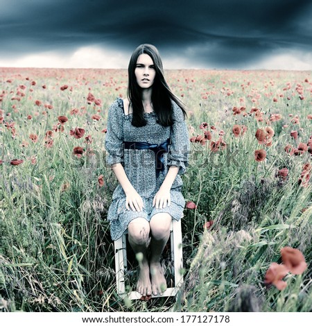 Mysterious portrait of young beautiful woman sitting on stool in a poppy field and looking at camera, summer nature outdoor. Toned, noise added.