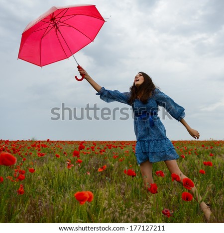 Young beautiful woman flying with wind holding red umbrella from a poppy field, summer outdoor.
