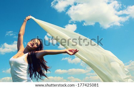 Happy young woman holding white scarf with opened arms expressing freedom, outdoor shot against blue sky, toned