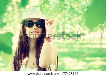 fashion girl outdoor portrait, young woman walking in summer park in sunglasses and fedora with long brown hair, toned and noise added