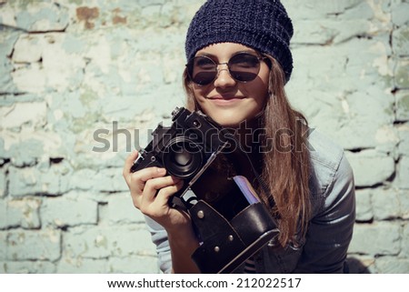 Retro photographer. Modern urban girl has fun with vintage photo camera outdoor near grunge wall, image toned.