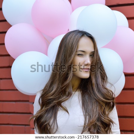 Happy young woman over red brick wall and holding pink and white balloons and gives a wink. Image toned.