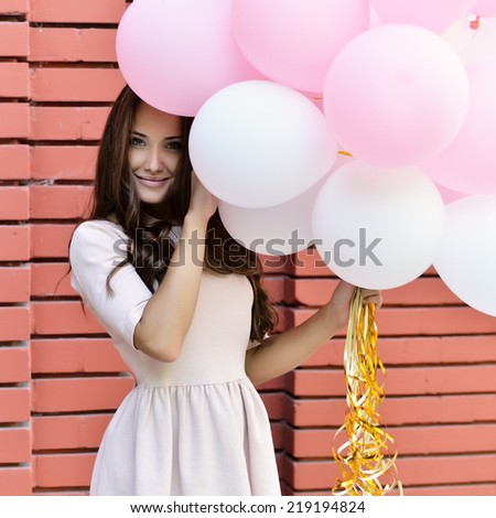 happy young woman standing over red brick wall and holding pink and white balloons