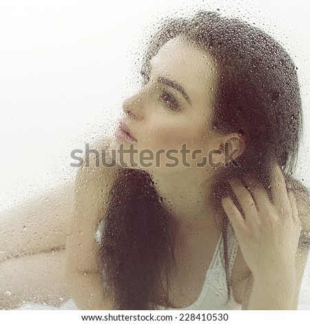 Beautiful young sensual woman is in bed on white sheet behind wet window, image toned.