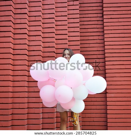 Happy young woman standing over red brick wall and holding pink and white balloons. Pleasure. Dreams.
