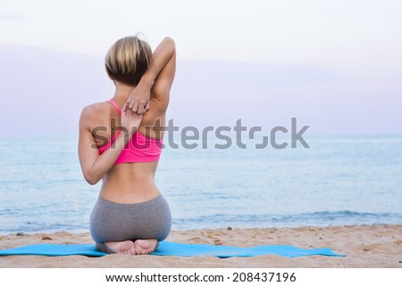 Stretching of muscles of the arms and back hands locking on the beach. Young beautiful girl on a rug on the sandy beach. Rays of the setting sun. Back view.