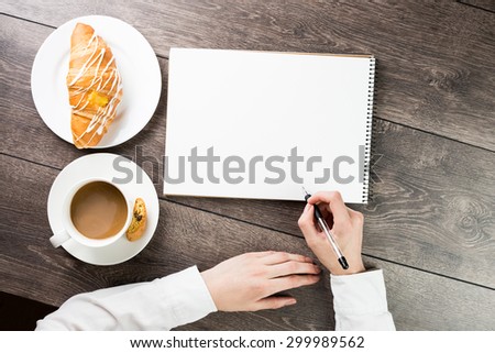 hand holding a pen. Top view. Table with notepads, cup of coffe and croissant . empty white space you can put your text.