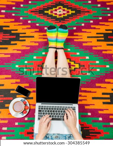 Laptop notebook computer phone mobile and tea cup in girl\'s hands sitting on a variegated colorful home rug. Black empty screen you can place your text information. Top view