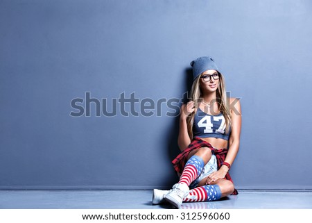 smiling beautiful young girl model in sport shirt, gray hat and Red checkered shirt, stylish glasses, red watch, white sneakers, leggings with USA flag, leaned on textured wall in the studio