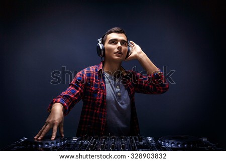 Closeup portrait of confident young DJ with stylish haircut and headphones on head mixing music on mixer looking up while standing isolated on dark background
