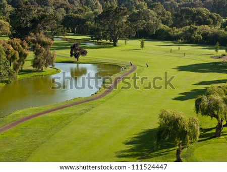 A high angle view of a golf course including a pond, cart path, and golfers.