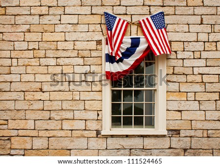 Red, white and blue bunting and crossed American flags adorn a casement style window set in a stand stone block wall.