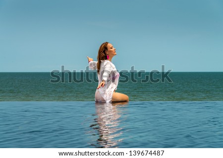 Woman in a wet white blouse and pink brassiere and earrings sitting on the edge of the pool and tans with closed eyes