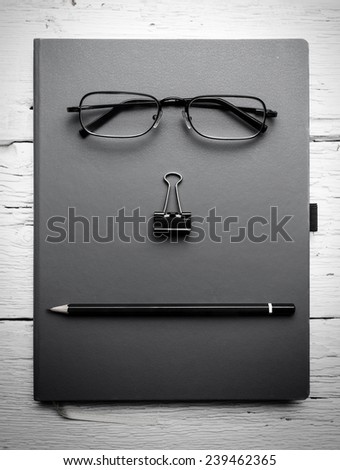 Notepad, eyeglasses, paperclip and pencil on wooden table. Black and white photo.