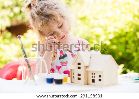 Portrait of little girl paints wooden model of house,  summer outdoor, new home concept