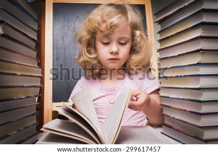 Portrait of cute smart girl reading a book while sitting with stack of books at table