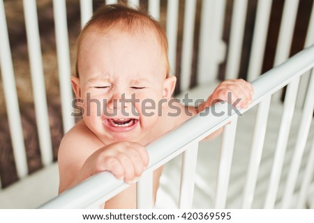 A year-old child is crying in the crib and holds onto the side of the bed
