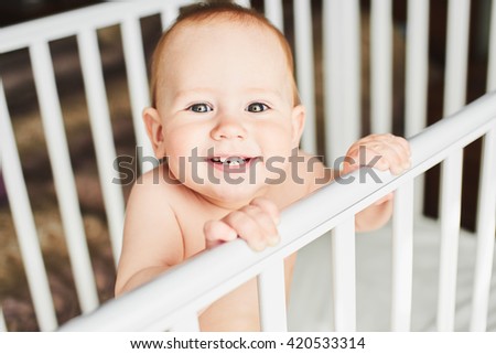 A year old baby is smiling in the crib and holds onto the side of the bed