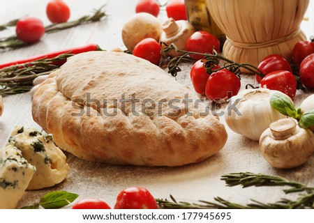 Calzone with ham mushrooms, meat, herbs, cook pours olive oil from the spoon. On the background, red wine, olive oil, cherry tomatoes, mushrooms and herbs.