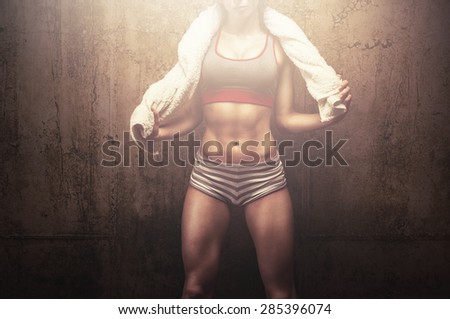 Exhausted fit young woman after hard workout using her soft white towel and proudly posing in front of gym textured background wall