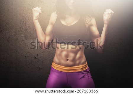 Fit young female fitness model posing and showing her muscular body with strong and tanned abdominal muscles in front of concrete wall