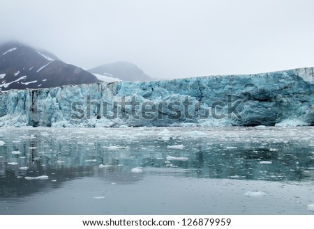 Amazing view of melting blue glacier, calm water and black mountain covered with snow against the background cloudy grey sky with heavy fog in Spitsbergen (Svalbard island), Greenland Sea, Norway