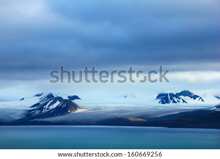Arctic scene - melting glacier, black mountains covered with snow and blue gulf against background of dramatic sky near Barentsburg, Spitsbergen archipelago (Svalbard island), Norway, Greenland sea