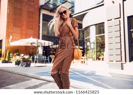 Fashionable woman walking in the street, wearing sunglasses, nice dress, high heels boots, handbag. Fashion urban autumn photo.