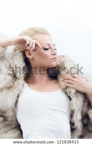 sexy blonde girl lying with open fur and withe singlet, her face is turned at left, her eyes are closed and the right hand is near the face