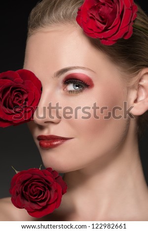 close-up portrait of blonde girl with three red roses near the face, she is turned of three quarters at right and looks in to the lens