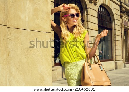 Beautiful blonde young woman wearing sunglasses, shorts, green top and handbag, standing on the street