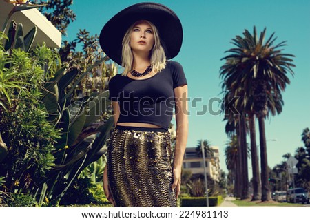 Beautiful blonde young woman wearing fashionable clothes, black hat walking on the street with palm trees. Fashion photo
