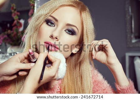 beautiful blonde young woman at beauty salon with smokey eyes. Make-up artist applying pink lipstick