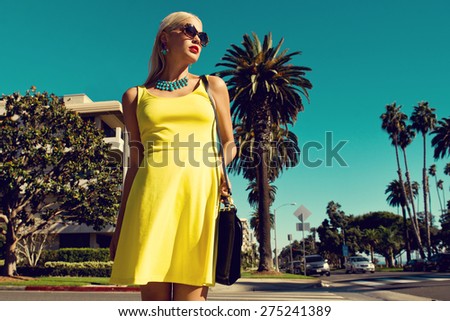 beautiful blonde woman posing with handbag in yellow dress and sunglasses walking across the street. Fashion photo