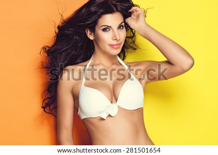 Sexy brunette woman wearing white bikini and colorful pants posing on bright yellow background. Perfect body. Skin care and cosmetics
