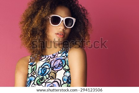 Beautiful woman posing in nice flower pattern dress and sunglasses on a pink background. Fashion photo with afro hairstyle.