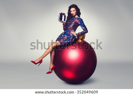 Christmas fashion photo of a nice woman sitting on a big red bauble holding a christmas gift