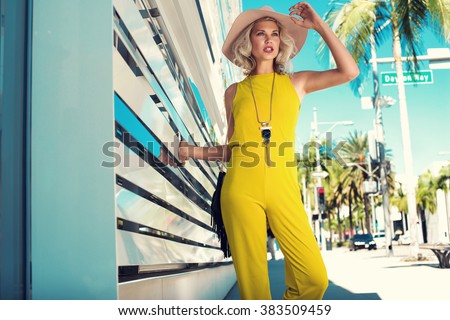 Sexy young woman walking in a city in yellow jumpsuit, nude hat, necklace, black handbag. Fashion summer photo. Bright colors