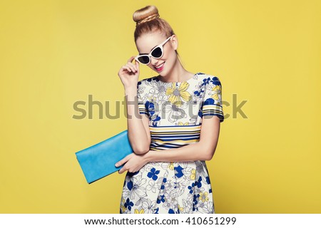 beautiful young blonde woman in nice spring dress, posing on yellow background in studio. Fashion photo, blue handbag and white sunglasses