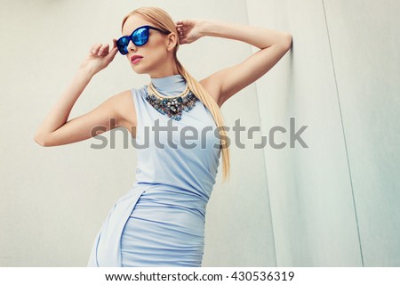 Beautiful young fashionable woman in sexy stylish dress, big necklace, blue sunglasses,  posing in the city.. Fashion photo