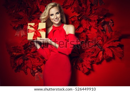 Beautiful young elegant woman in sexy red dress posing over red background with poinsettia holding present. Christmas photo