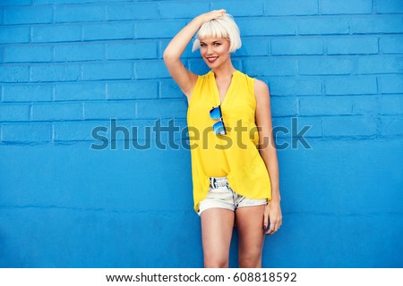 Beautiful young woman walking in a city next to blue wall in jeans shorts, yellow blouse and sunglasses. Fashion summer photo. Big smile