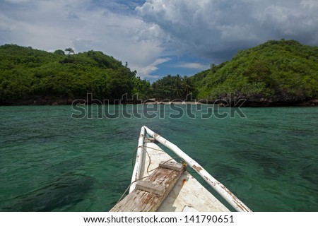 White painted prow of an outrigger canoe sailing across emerald green ocean water toward the sandy beach of a remote tropical Philippine Island in Southeast Asia.
