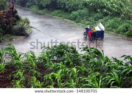 A Filipino woman pushing her bike with sidecar loaded with large sacks up a road in a heavy rain storm somewhere in the Philippine Islands.