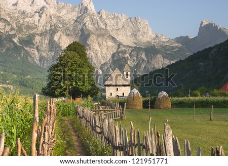 a path with wooden fence leads the catholic church in the Theth Valley, on background the mountains of Albanian Alps
