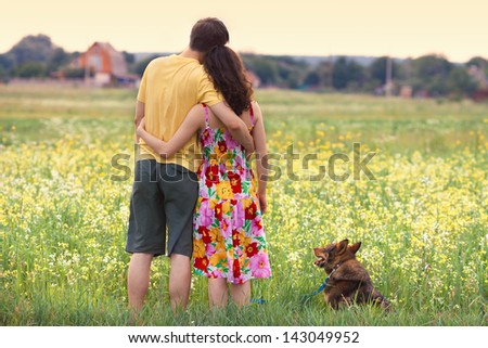 Young Couple Hugging In The Field And Looking At The Village At Sunset. Dog Sitting Near In The Meadow