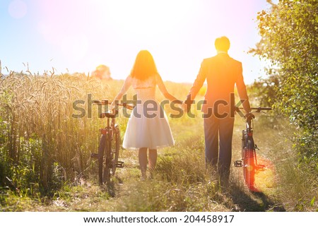Young happy bride and groom carry bikes in the field back to camera and holding hands.